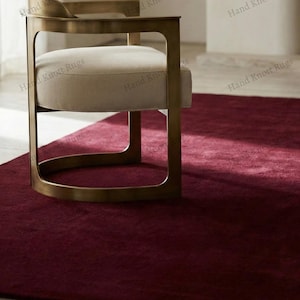 May include: A burgundy-colored rug with a soft texture. A modern chair with a gold-colored metal frame and a cream-colored seat sits on the rug. The rug fills the foreground, with the chair in the background.