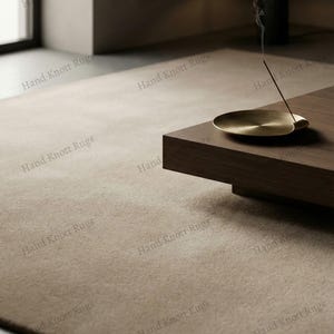 Puede incluir: Una alfombra beige texturizada llena la imagen, complementada con una mesa de centro de madera oscura. Un quemador de incienso de latón con una varilla de incienso encendida está sobre la mesa, con humo ascendente. La escena evoca un diseño interior minimalista.
