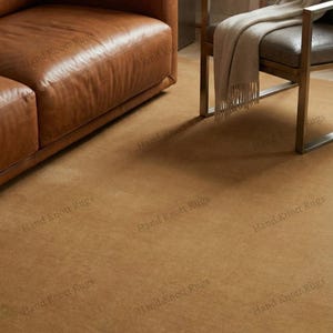May include: A warm-toned, solid-colored rug fills the frame, complementing the leather furniture and chair. The rug's texture is visible, adding depth to the room's design. The rug is a neutral color, suitable for various interior styles.