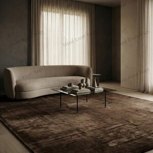 May include: A large, brown area rug with a distressed, textured appearance. The rug is placed in a living room setting, with a curved beige sofa, a glass-top coffee table, and neutral-toned curtains. The text "Hand Knott Rugs" is visible.