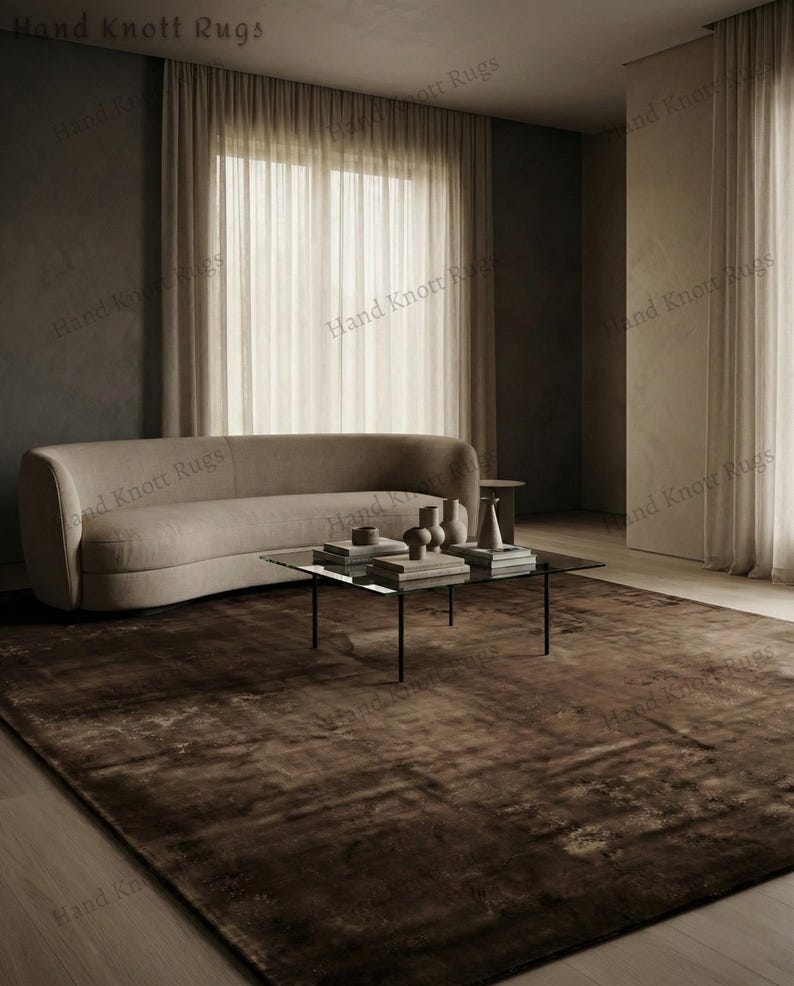 May include: A large, brown area rug with a distressed, textured appearance. The rug is placed in a living room setting, with a curved beige sofa, a glass-top coffee table, and neutral-toned curtains. The text "Hand Knott Rugs" is visible.