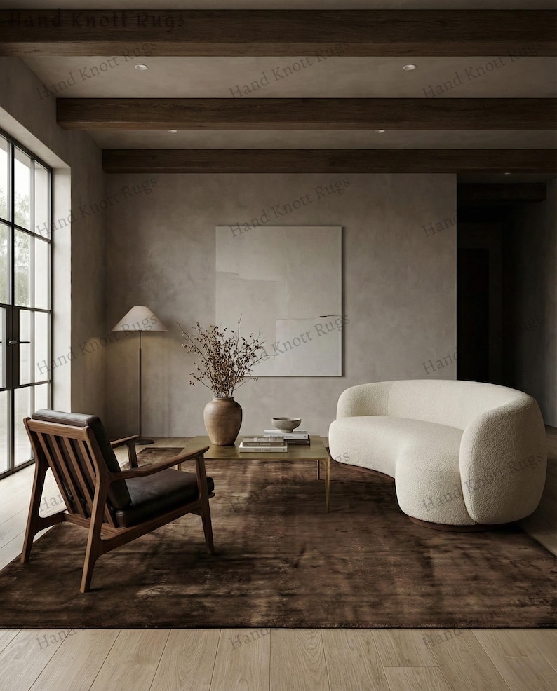 May include: A modern living room featuring a dark brown rug, a curved white sofa, and a wooden armchair with a leather seat. A coffee table with a vase of dried flowers and a lamp are also present.