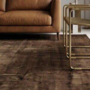 May include: A brown area rug with a distressed, textured appearance. The rug is placed beneath a brown leather sofa and a gold-colored side table. The rug's design features a mottled pattern, adding visual interest to the room.