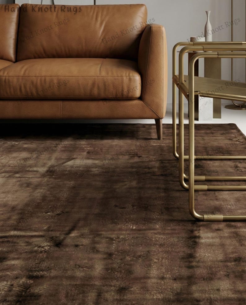 May include: A brown area rug with a distressed, textured appearance. The rug is placed beneath a brown leather sofa and a gold-colored side table. The rug's design features a mottled pattern, adding visual interest to the room.