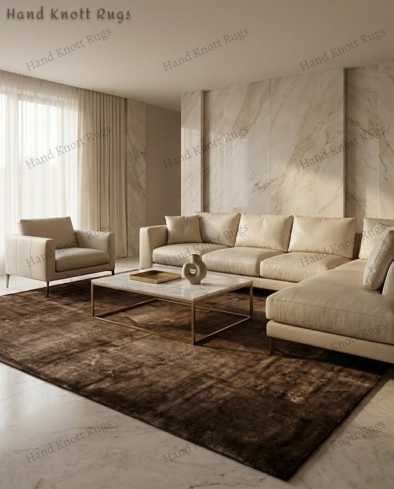 May include: A modern living room featuring a large, brown area rug. The room includes a beige sectional sofa, a matching armchair, and a marble-topped coffee table with a gold frame. The walls are a light beige color.