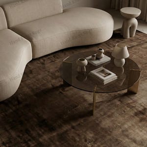 May include: A modern living room setup featuring a beige curved sofa, a round glass coffee table with gold legs, and a textured brown rug. Decorative items include vases, books, and a lamp. The text "Hand Knott Rugs" is visible.