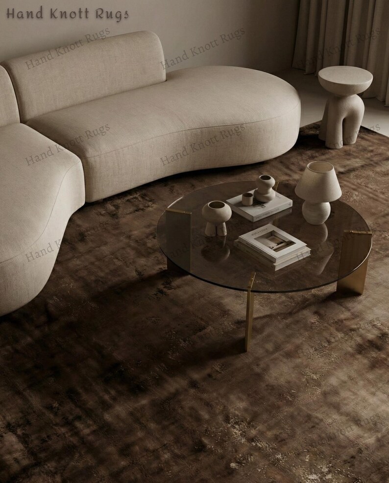 May include: A modern living room setup featuring a beige curved sofa, a round glass coffee table with gold legs, and a textured brown rug. Decorative items include vases, books, and a lamp. The text "Hand Knott Rugs" is visible.