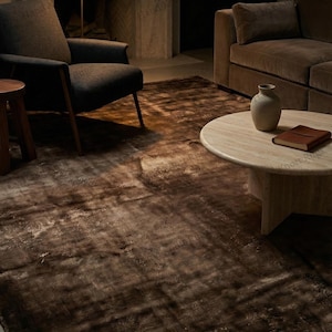 May include: A brown area rug with a distressed, mottled appearance. The rug is placed in a living room setting, with a dark armchair, a round coffee table, and a brown sofa visible in the background. The rug's texture adds warmth to the space.