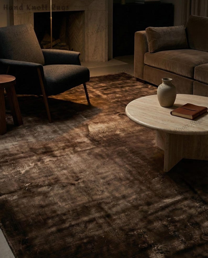 May include: A brown area rug with a distressed, mottled appearance. The rug is placed in a living room setting, with a dark armchair, a round coffee table, and a brown sofa visible in the background. The rug's texture adds warmth to the space.