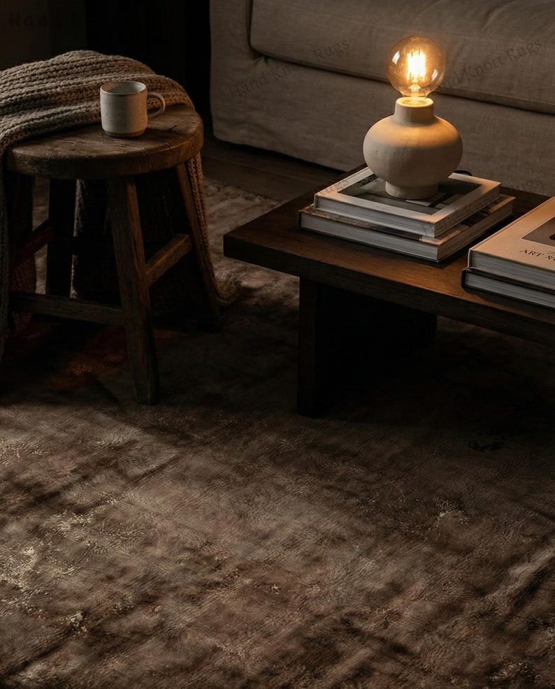 May include: A cozy living room scene featuring a dark brown rug, a wooden side table with a mug and blanket, and a coffee table with books and a lamp. The lamp has a beige base and a warm light bulb.