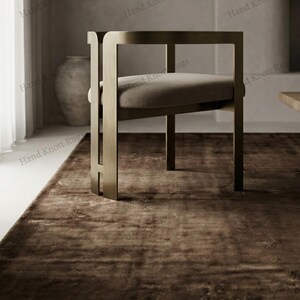 May include: A modern chair with a rectangular metal frame and a cushioned seat sits on a brown rug. The rug has a distressed, textured appearance. The words "Hand Knott Rugs" are printed on the top and sides of the image.