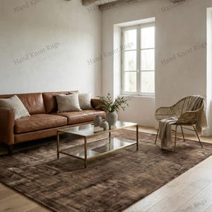 May include: A brown area rug with a distressed pattern in a living room setting. The rug is placed under a glass-top coffee table and a brown leather sofa. The room has a neutral color palette with natural light.