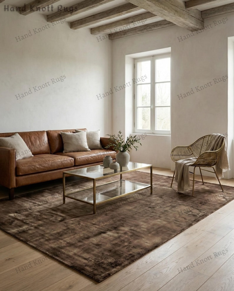 May include: A brown area rug with a distressed pattern in a living room setting. The rug is placed under a glass-top coffee table and a brown leather sofa. The room has a neutral color palette with natural light.