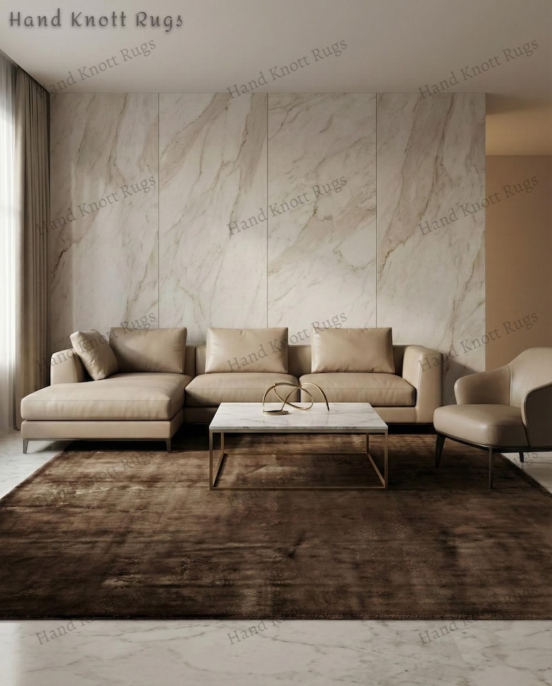 May include: A modern living room featuring a large, brown rug. A beige sectional sofa and a matching armchair are arranged around a marble-topped coffee table with a gold base. The wall is a light marble pattern.