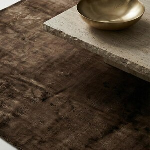 May include: A close-up of a brown rug with a textured surface. A gold-colored bowl sits on a light-colored stone table. The rug and table create a modern, minimalist aesthetic.