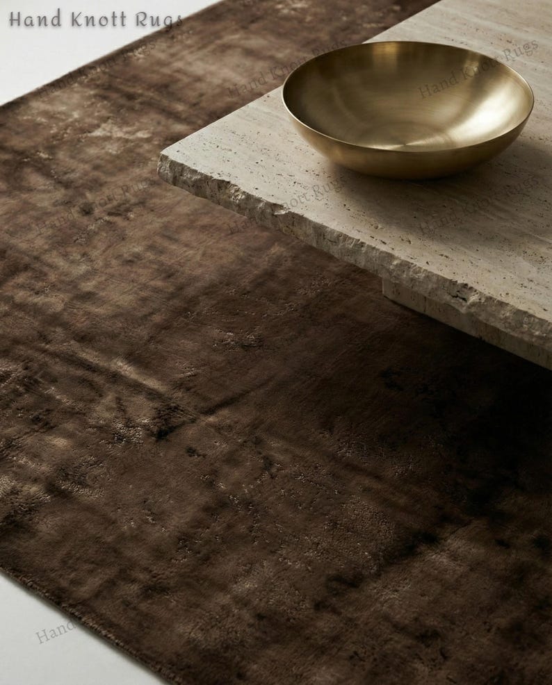 May include: A close-up of a brown rug with a textured surface. A gold-colored bowl sits on a light-colored stone table. The rug and table create a modern, minimalist aesthetic.