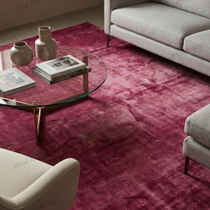 Pode incluir: Um tapete magenta vibrante e texturizado domina a cena, complementado por um sofá e cadeiras cinzentos modernos. Uma mesa de centro com tampo de vidro e pernas douradas exibe livros e vasos decorativos. A cor do tapete adiciona calor.