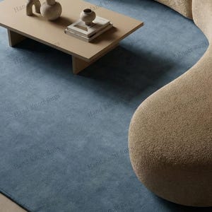 Könnte beinhalten: Ein modernes Wohnzimmer mit einem hellblauen Teppich, einem beigen Couchtisch mit dekorativen Vasen und Büchern sowie einem geschwungenen beigen Sofa. Der Teppich bedeckt den Großteil des Bodens und schafft ein stimmiges und stilvolles Interieur.