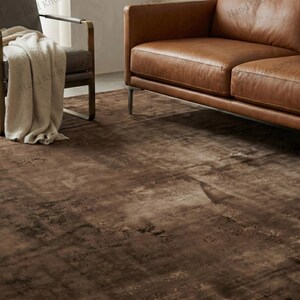 May include: A brown area rug with a distressed, textured appearance. The rug is the focal point of a living room setting, with a leather sofa and armchair visible in the background. The rug's color palette includes shades of brown and tan.