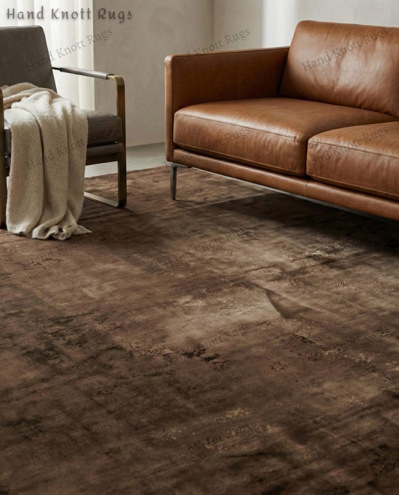 May include: A brown area rug with a distressed, textured appearance. The rug is the focal point of a living room setting, with a leather sofa and armchair visible in the background. The rug's color palette includes shades of brown and tan.