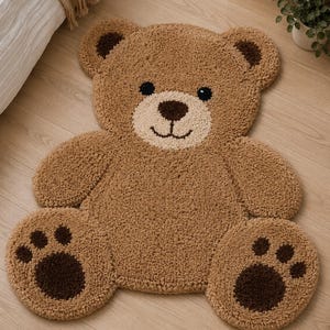 Pode incluir: Um tapete castanho em forma de urso de pelúcia com rosto castanho claro e detalhes castanhos escuros. O tapete tem uma textura fofa e está colocado sobre um chão de madeira clara. Os olhos e o nariz do urso são castanhos escuros.