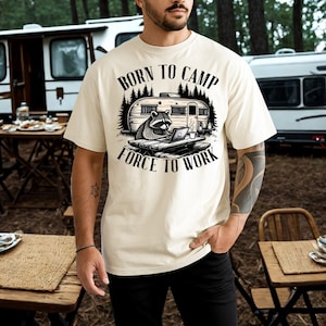 May include: A cream-colored short-sleeve t-shirt featuring a black graphic of a raccoon sitting at a picnic table with a laptop in front of a camper. The text above reads BORN TO CAMP and the text below reads FORCE TO WORK in a bold font.
