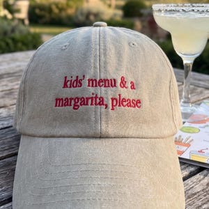 May include: A beige baseball cap featuring the embroidered red text "kids' menu & a margarita, please" centered on the front panel. The cap has a structured crown, a curved brim, and stitched eyelets for ventilation on the top panels.
