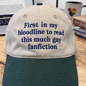 May include: A beige baseball cap with a dark green brim. The front of the cap features embroidered dark blue text that reads: First in my bloodline to read this much gay fanfiction. The cap has a small dark green button at the top center.