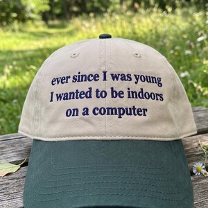 May include: A two-tone baseball cap featuring a beige crown and a dark green brim. The front of the crown displays the embroidered text: ever since I was young I wanted to be indoors on a computer. The cap has a small dark button at the top center.