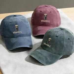 May include: Three baseball caps in shades of blue, burgundy, and green. Each cap features a shamrock and beer glass graphic with the text "Vitamin G". The caps are made of a soft, worn fabric.