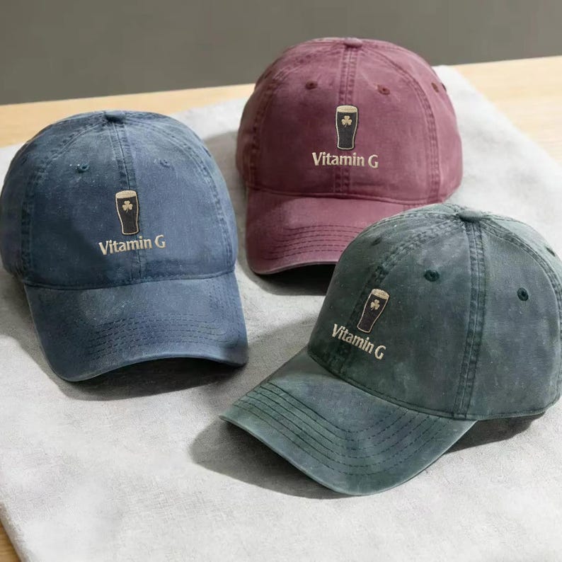 May include: Three baseball caps in shades of blue, burgundy, and green. Each cap features a shamrock and beer glass graphic with the text "Vitamin G". The caps are made of a soft, worn fabric.