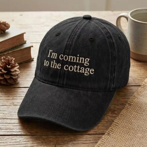 May include: Black baseball cap with the embroidered text "I'm coming to the cottage" in a casual font. The cap is made of a soft, worn fabric and has a curved brim. A mug and books are in the background.