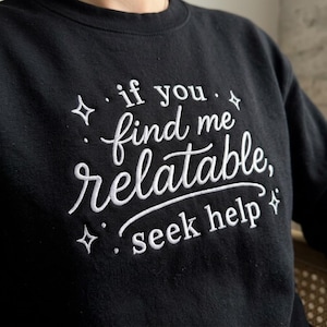 Pu&ograve; includere: Una felpa nera a girocollo con testo ricamato in bianco che recita "if you find me relatable, seek help", circondata da piccoli accenti a forma di stella ricamati. La scritta utilizza un mix di caratteri corsivi e serif.