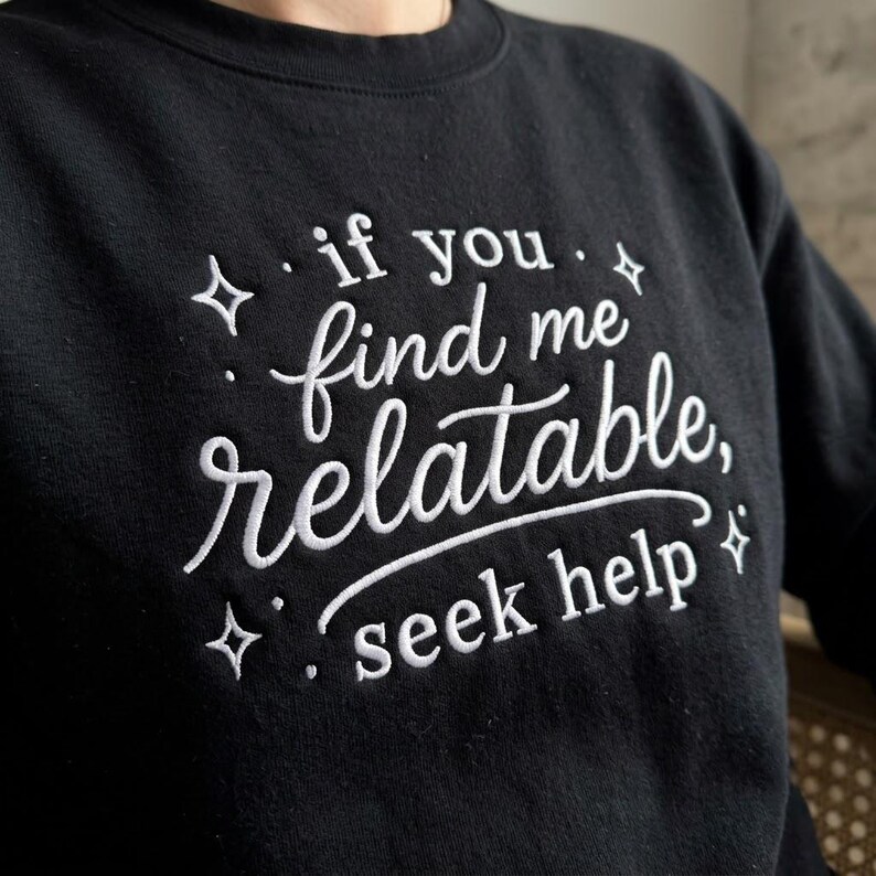 Pu&ograve; includere: Una felpa nera a girocollo con testo ricamato in bianco che recita "if you find me relatable, seek help", circondata da piccoli accenti a forma di stella ricamati. La scritta utilizza un mix di caratteri corsivi e serif.