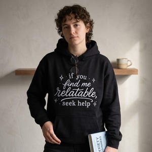 Pu&ograve; includere: Una felpa nera con cappuccio che presenta il testo bianco "if you find me relatable, seek help" circondato da piccole icone a forma di stella. La felpa include una tasca a marsupio frontale e maniche lunghe, con occhiali appesi al collo.