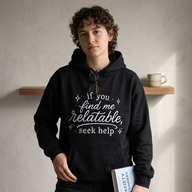 Pu&ograve; includere: Una felpa nera con cappuccio che presenta il testo bianco "if you find me relatable, seek help" circondato da piccole icone a forma di stella. La felpa include una tasca a marsupio frontale e maniche lunghe, con occhiali appesi al collo.