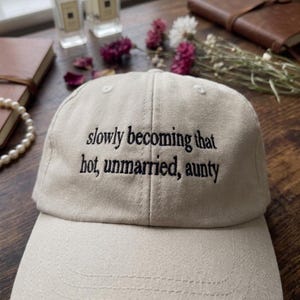 May include: A beige baseball cap featuring black embroidered text on the front that reads, slowly becoming that hot, unmarried, aunty. The cap has a structured crown with a curved brim and visible stitching details across the fabric surface.