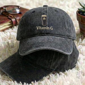 May include: A dark gray baseball cap with the text "Vitamin G" and a graphic of a pint of beer with a shamrock. The cap is made of a worn, textured fabric and has a curved brim. The cap is sitting on top of another cap.