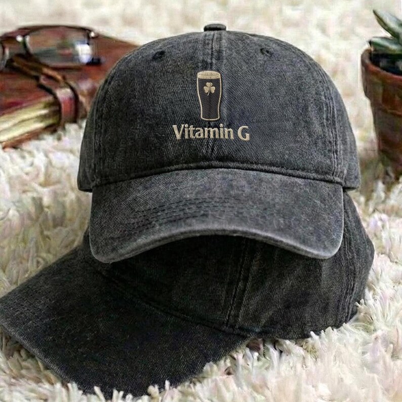 May include: A dark gray baseball cap with the text "Vitamin G" and a graphic of a pint of beer with a shamrock. The cap is made of a worn, textured fabric and has a curved brim. The cap is sitting on top of another cap.