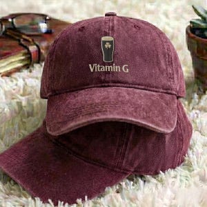 May include: A maroon baseball cap with a curved brim. The cap features an embroidered design of a pint of beer with a shamrock and the text "Vitamin G". The hat is made of a soft, worn material.