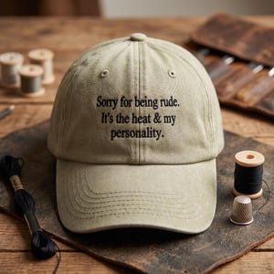 May include: A beige baseball cap featuring black embroidered text that reads, Sorry for being rude. It's the heat & my personality. The cap has a structured crown, a curved brim, and stitched eyelets on the top panels for ventilation.