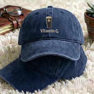 May include: Two navy blue baseball caps are stacked on top of each other. The top cap features a graphic of a pint of beer with a shamrock and the words "Vitamin G" in a gold color.