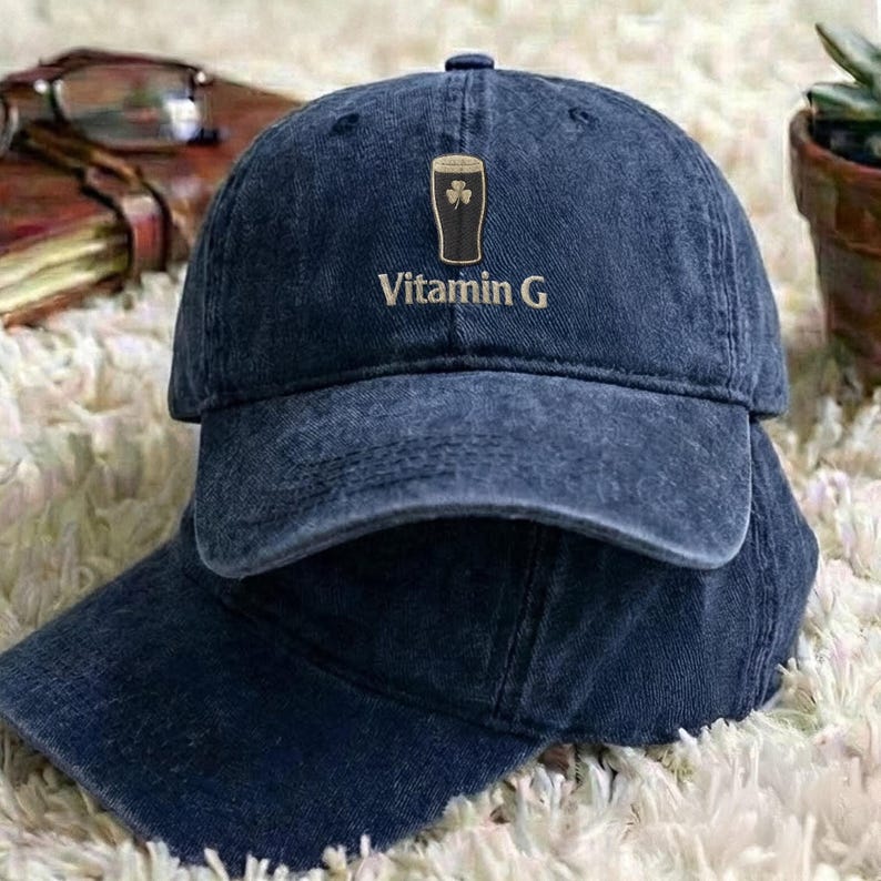 May include: Two navy blue baseball caps are stacked on top of each other. The top cap features a graphic of a pint of beer with a shamrock and the words "Vitamin G" in a gold color.