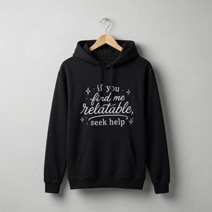 Pu&ograve; includere: Una felpa con cappuccio nera con la scritta bianca "if you find me relatable, seek help" circondata da piccole icone a forma di stella. Il capo presenta una tasca a marsupio frontale, maniche lunghe e un cappuccio con coulisse, esposto su una gruccia.
