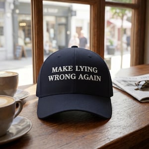 Może przedstawiać: Granatowa czapka z daszkiem z napisem "MAKE LYING WRONG AGAIN" w białych literach. Czapka leży na drewnianej powierzchni, w pobliżu dwóch filiżanek kawy. W tle widoczna jest gazeta i okulary przeciwsłoneczne.