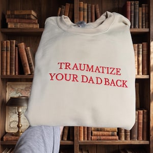 May include: A beige crewneck sweatshirt featuring the embroidered red text TRAUMATIZE YOUR DAD BACK centered on the chest. The garment has long sleeves, a ribbed collar, and a standard fit, presented in a clear, straightforward design.