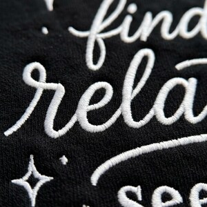 Pu&ograve; includere: Una superficie in tessuto nero con testo ricamato in bianco in corsivo che recita find, rela e see. Il ricamo include piccoli accenti a forma di stella a quattro punte sparsi tra le righe di testo, creando un aspetto strutturato e in rilievo.
