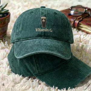 May include: A dark green baseball cap with a curved brim. The cap features an embroidered design of a pint of beer with a shamrock and the text "Vitamin G" in white. The cap is made of a soft, worn material.
