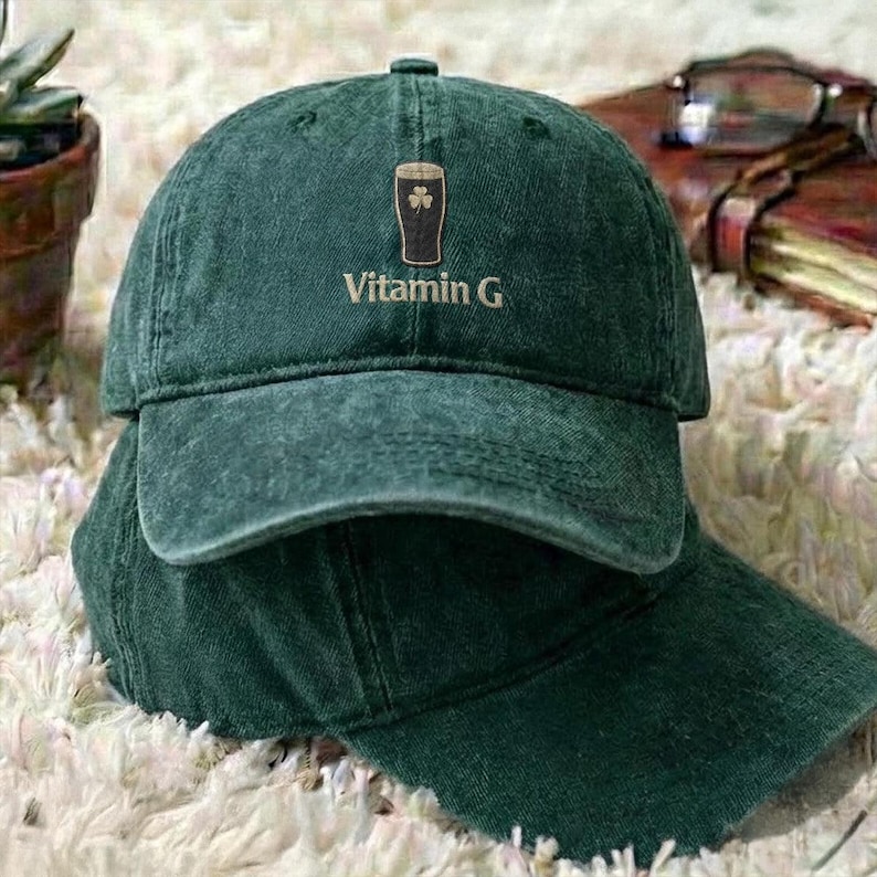 May include: A dark green baseball cap with a curved brim. The cap features an embroidered design of a pint of beer with a shamrock and the text "Vitamin G" in white. The cap is made of a soft, worn material.