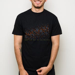 May include: Black t-shirt with a binary code design in orange, blue, and white. The text "ANOTHER DAY IN THE MATRIX" is printed in green at the bottom of the design. The shirt is a crew neck style.